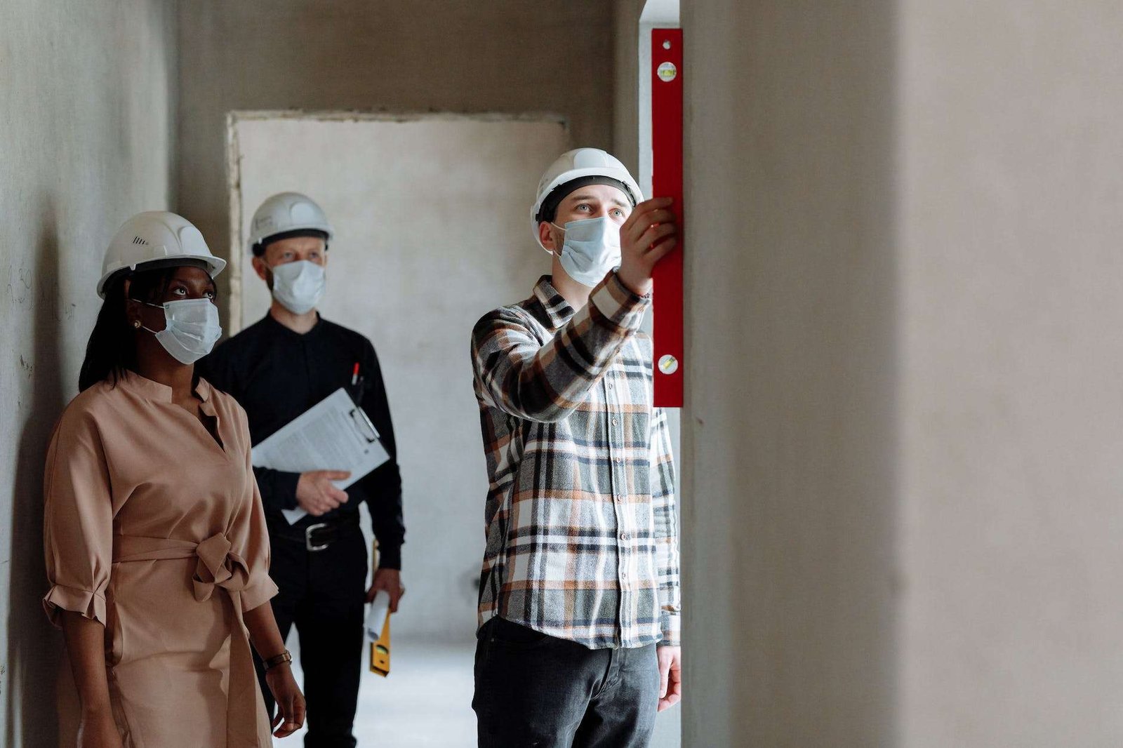 a man and woman wearing face mask while looking at the man in plaid long sleeves measuring the wall