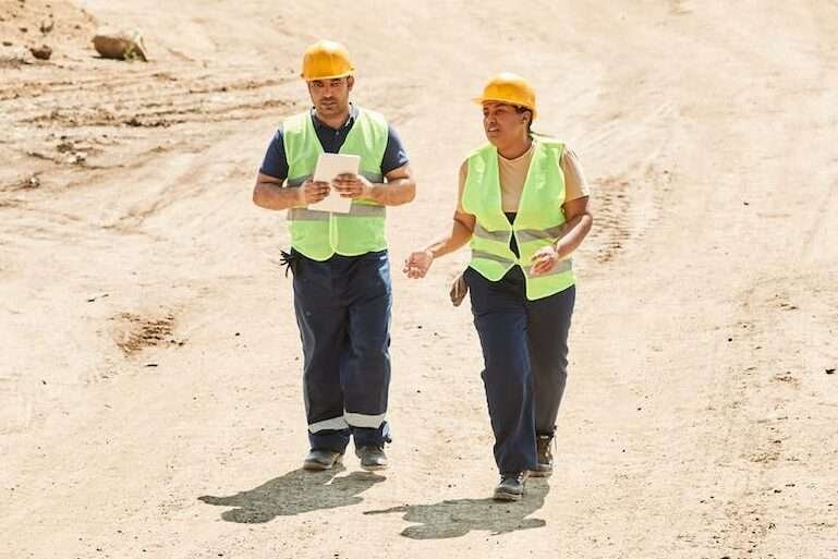 man and woman in helmets and reflective vests walking on a construction site